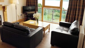 A living room with two black sofas, a coffee table, a TV, and a view of a lake through large windows.