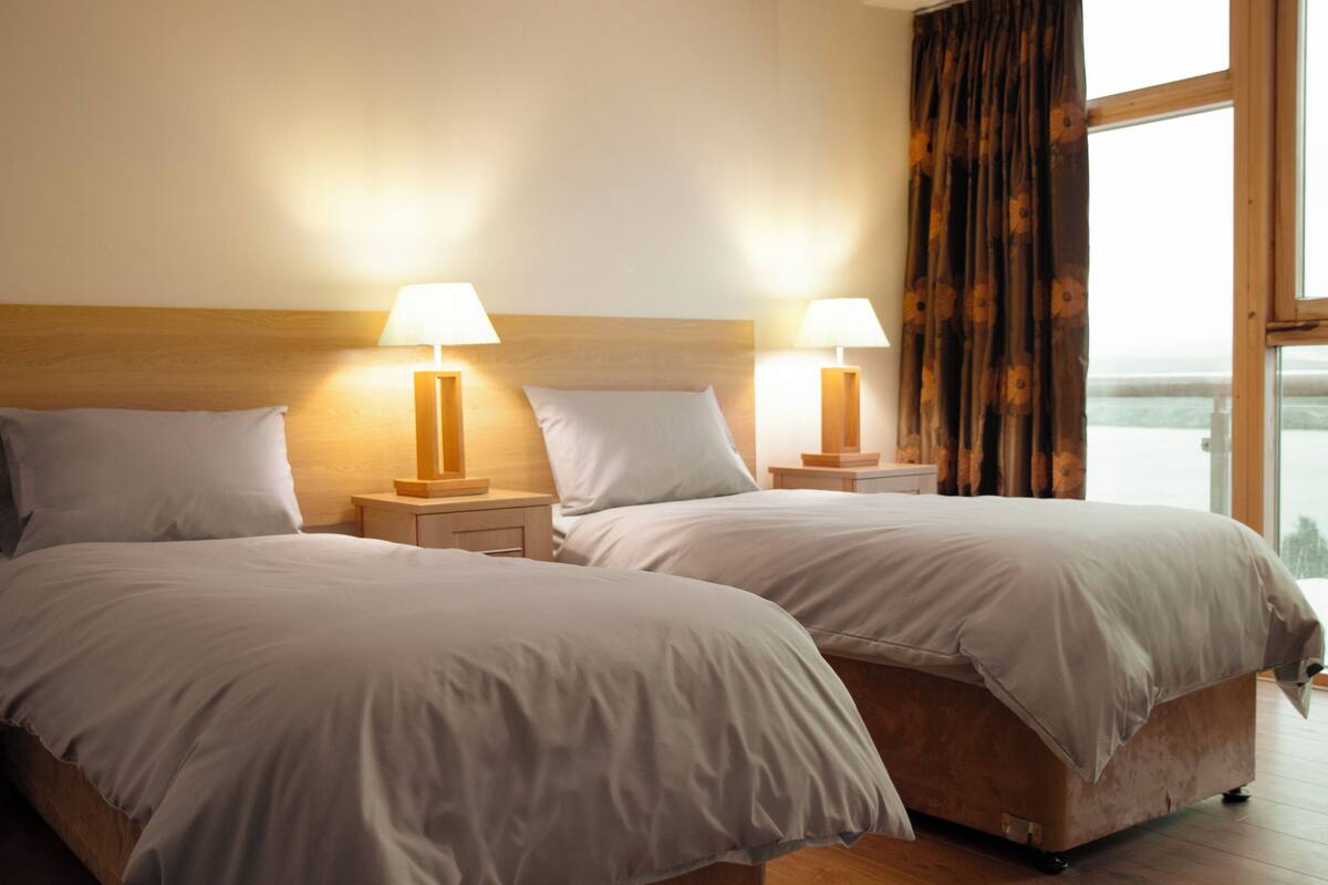 Una habitació d'hotel amb dos llits, cadascun amb una tauleta de nit i una làmpada. L'habitació té una finestra gran amb cortines, i les parets estan pintades de color clar.
