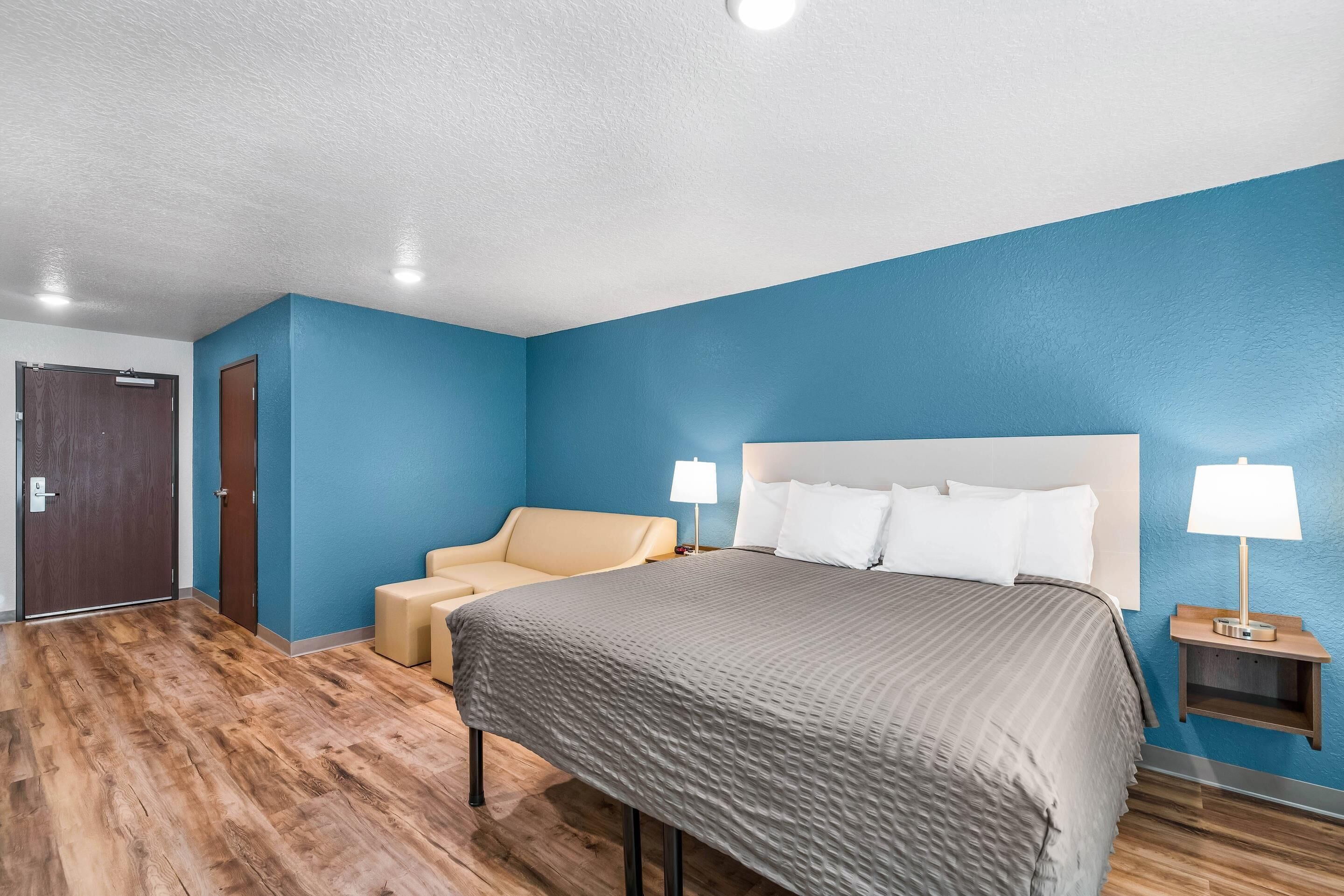 A hotel room with a large bed, a chair, a nightstand with a lamp, and a wooden floor.