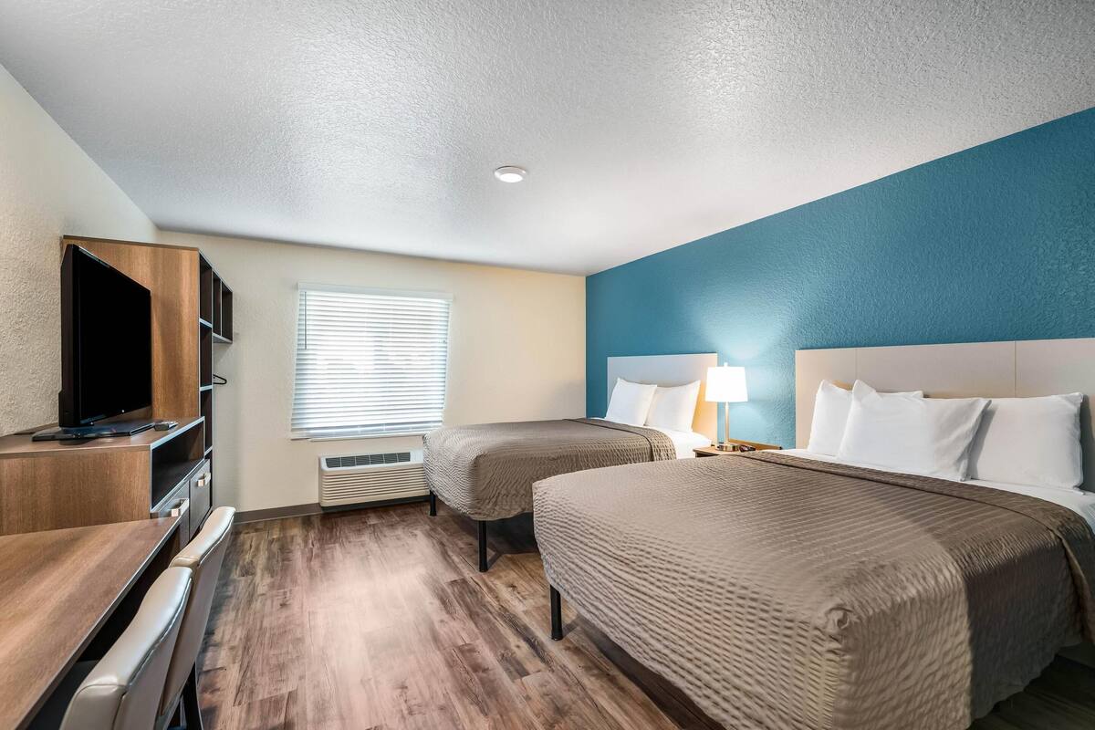 A hotel room with two beds, a TV, a desk, and a window with blinds.