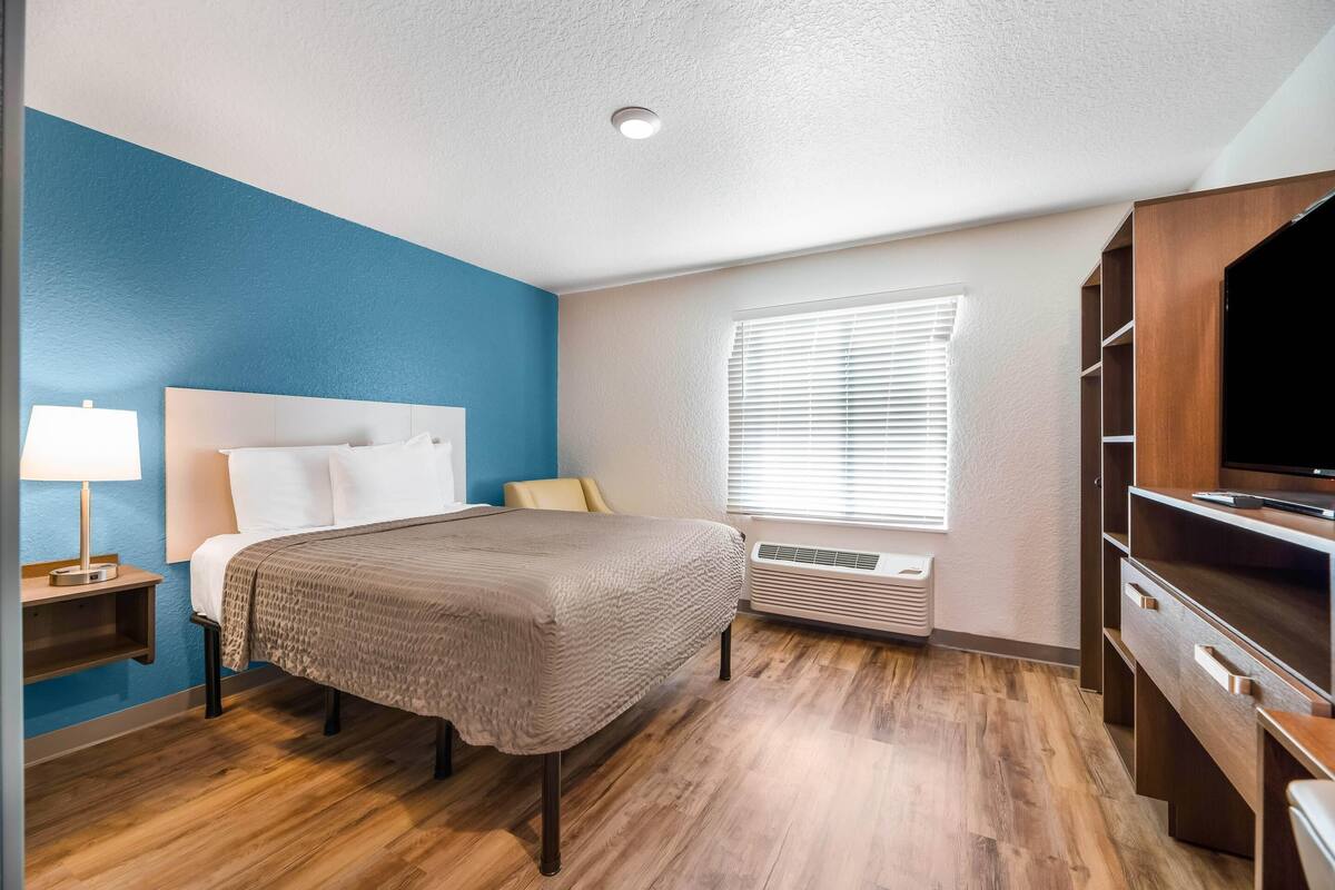A hotel room with a bed, bedside table, lamp, air conditioning unit, window with blinds, and a TV stand.