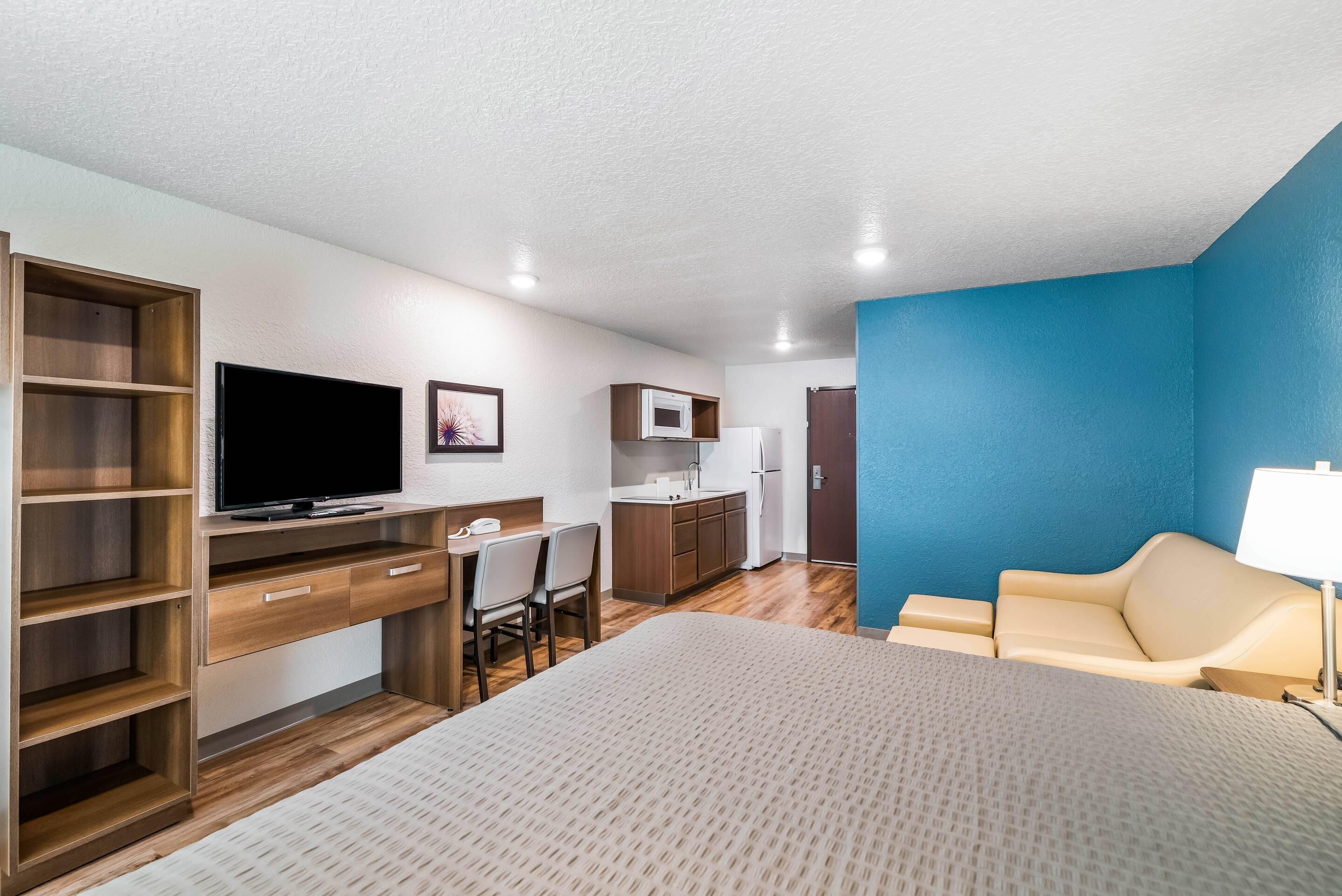 Una habitación de hotel compacta con cama, un escritorio y silla, televisión, kitchenette y una pared de acento azul.