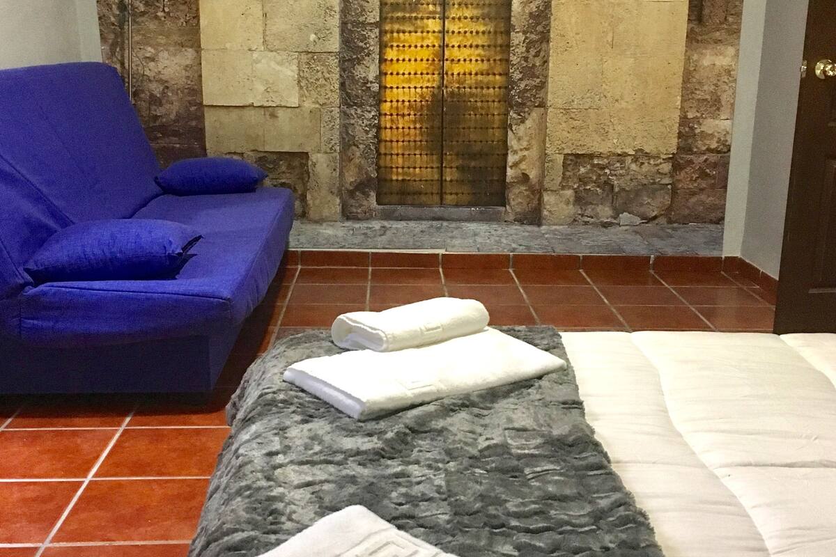 Um quarto com parede de pedra, um sofá azul, uma cama com colchão branco e duas toalhas sobre um cobertor.