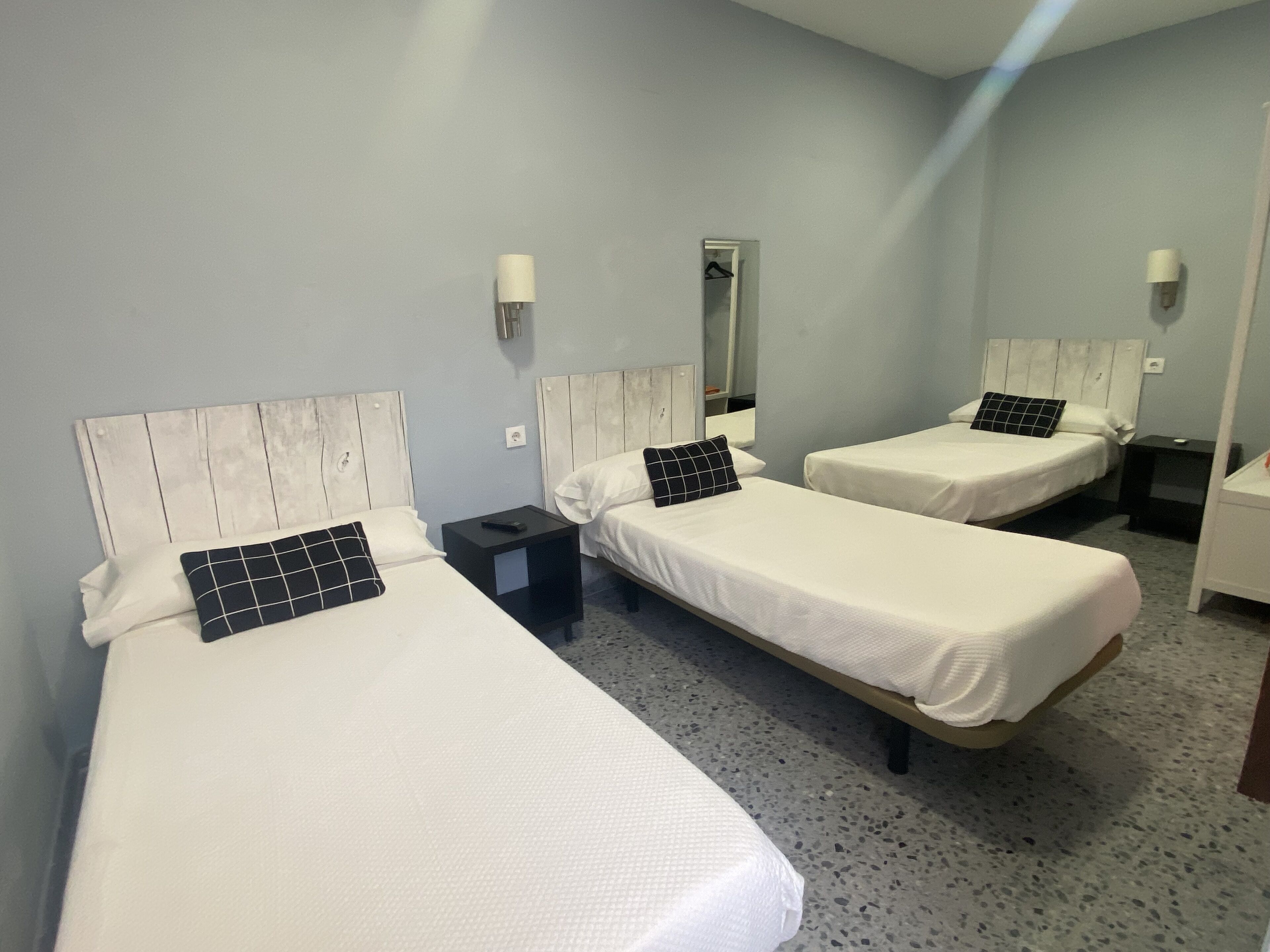 Ett hotellrum med tre enkelsängar, var och en med en vit madrass och en svartvit kudde. Det finns sängbord med lampor, en spegel på väggen och ett litet sidobord.