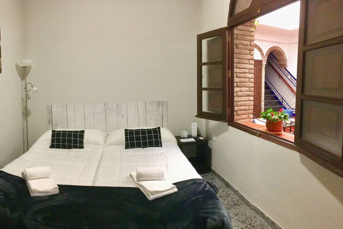 Quarto de hotel com cama, mesa de cabeceira, janela com vista para um pátio e um quadro na parede.