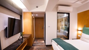 Habitació d'hotel amb un llit, una televisió penjada a la paret, un armari de fusta i un bany visible a través d'una porta de vidre.