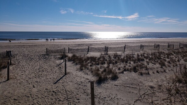 En strand med ett trästaket, sandig mark och havet i bakgrunden.