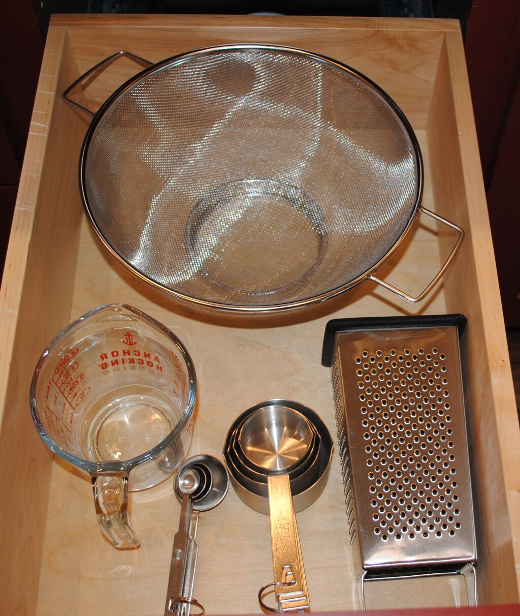A wooden tray with a metal strainer, a measuring cup, a small metal bowl, a grater, and a metal measuring spoon.