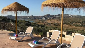 En terrass med vita loungestolar och parasoller med halmtak, med utsikt över ett vackert landskap.