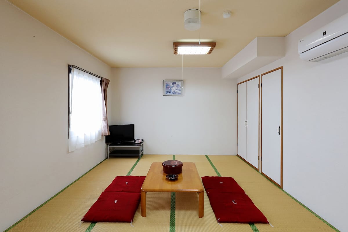 A room with tatami mat flooring, a wooden table, two red cushions, a black chair, a ceiling light, an air conditioner, and a framed picture on the wall.