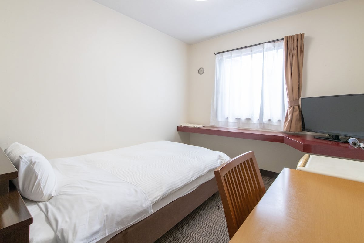 A small hotel room with a single bed, a wooden table, a TV, and a window with curtains.