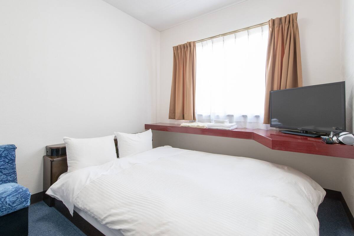 A hotel room with a bed, a TV, a desk with a computer, and a window with curtains.