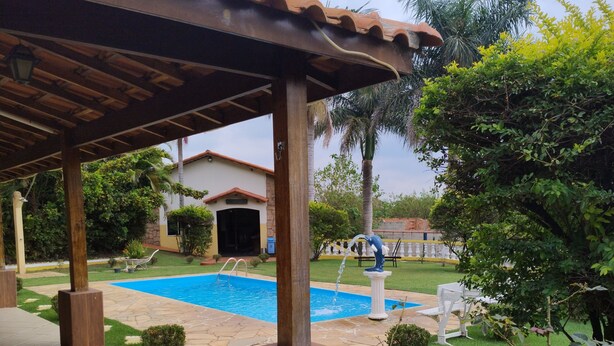 A covered patio area with a swimming pool, surrounded by greenery and a white building in the background. - Chácara Recanto do baiano (Sorocaba)