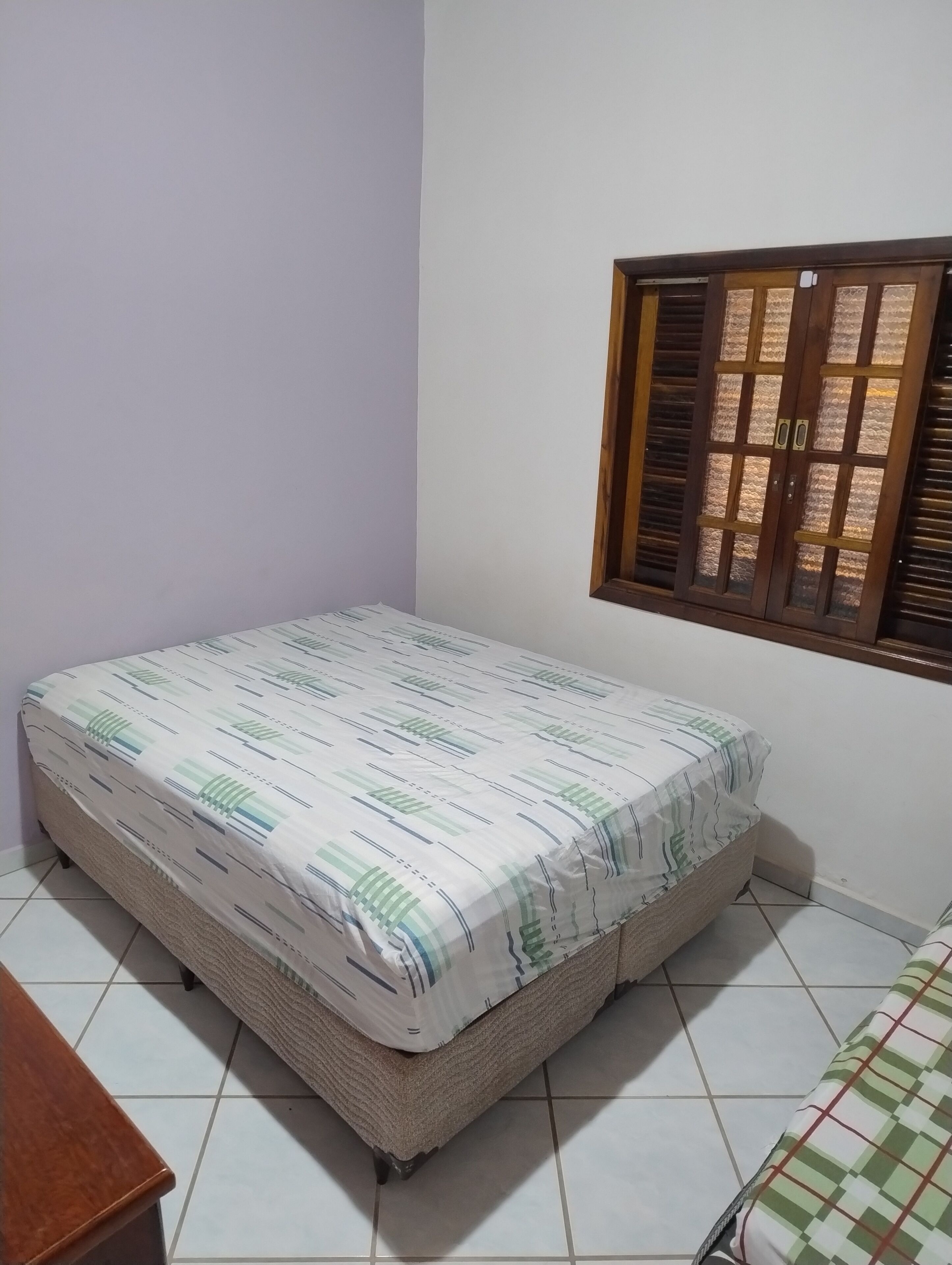 A small, neatly made bed with a patterned bedspread in a room with white walls and a window with wooden shutters.