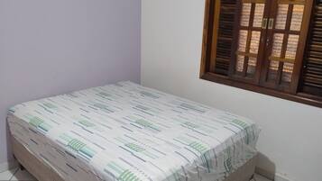 A small, neatly made bed with a patterned bedspread in a room with white walls and a window with wooden shutters.