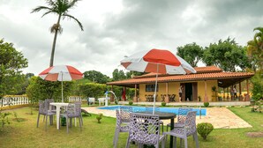A poolside area with outdoor seating and umbrellas, a pool, and a building with a tiled roof. - Chácara Recanto do baiano (Sorocaba)