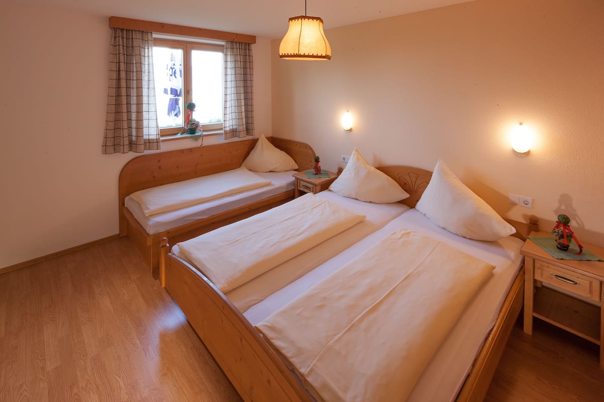 Une chambre avec deux lits en bois, une fenêtre avec des rideaux, une table de chevet avec une lampe et un luminaire fixé au mur.