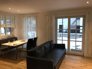 A modern living room with a black sofa, a dining table with chairs, and a large window with curtains. - Ferienwohnung Lea am See (Tegernsee)