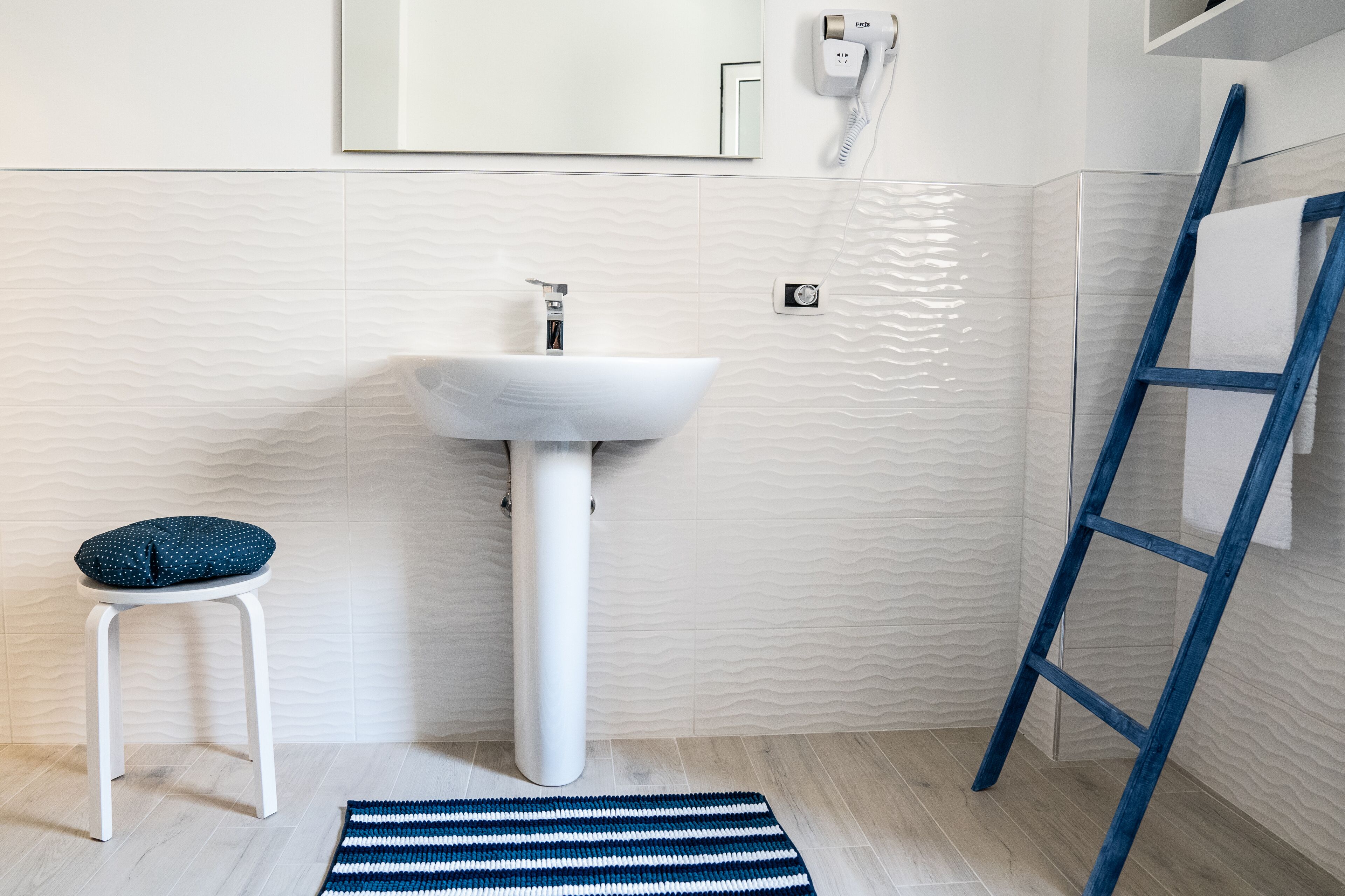 A bathroom with a white sink, a blue stool, a blue and white striped rug, and a blue ladder leaning against the wall.