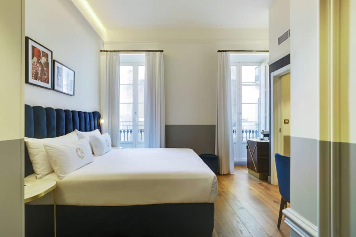 A hotel room with a large bed, a bedside table, a wardrobe, and a view of the city through the windows.