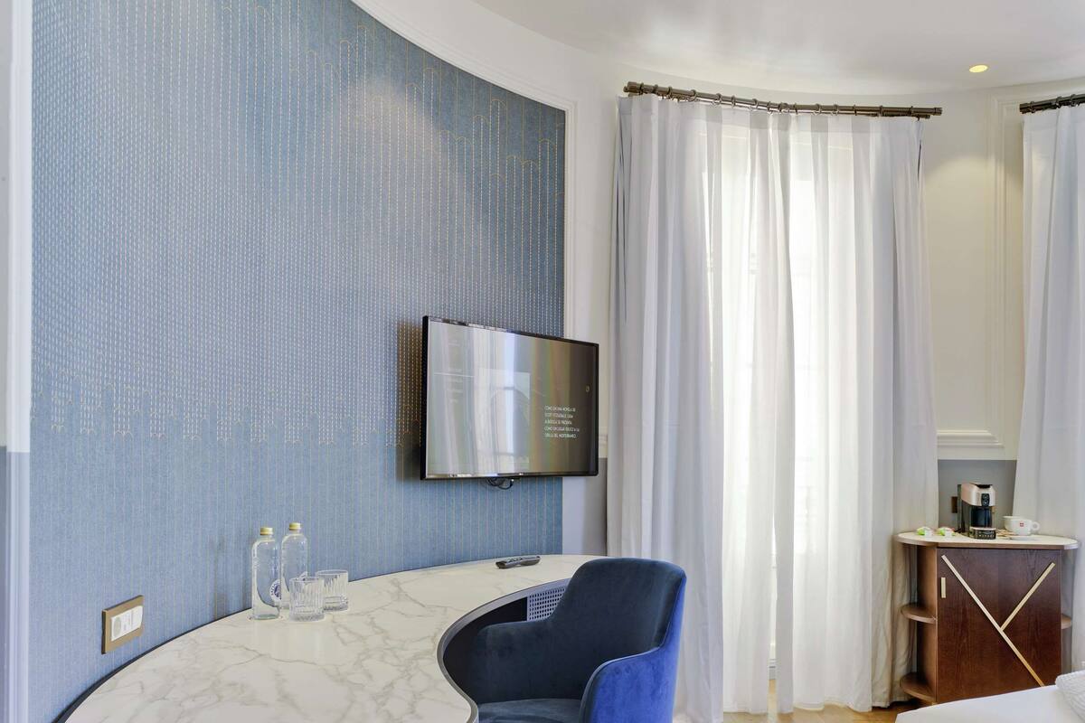 A hotel room with a curved marble table, a blue chair, a bed, a TV mounted on the wall, and a small desk with a coffee maker.