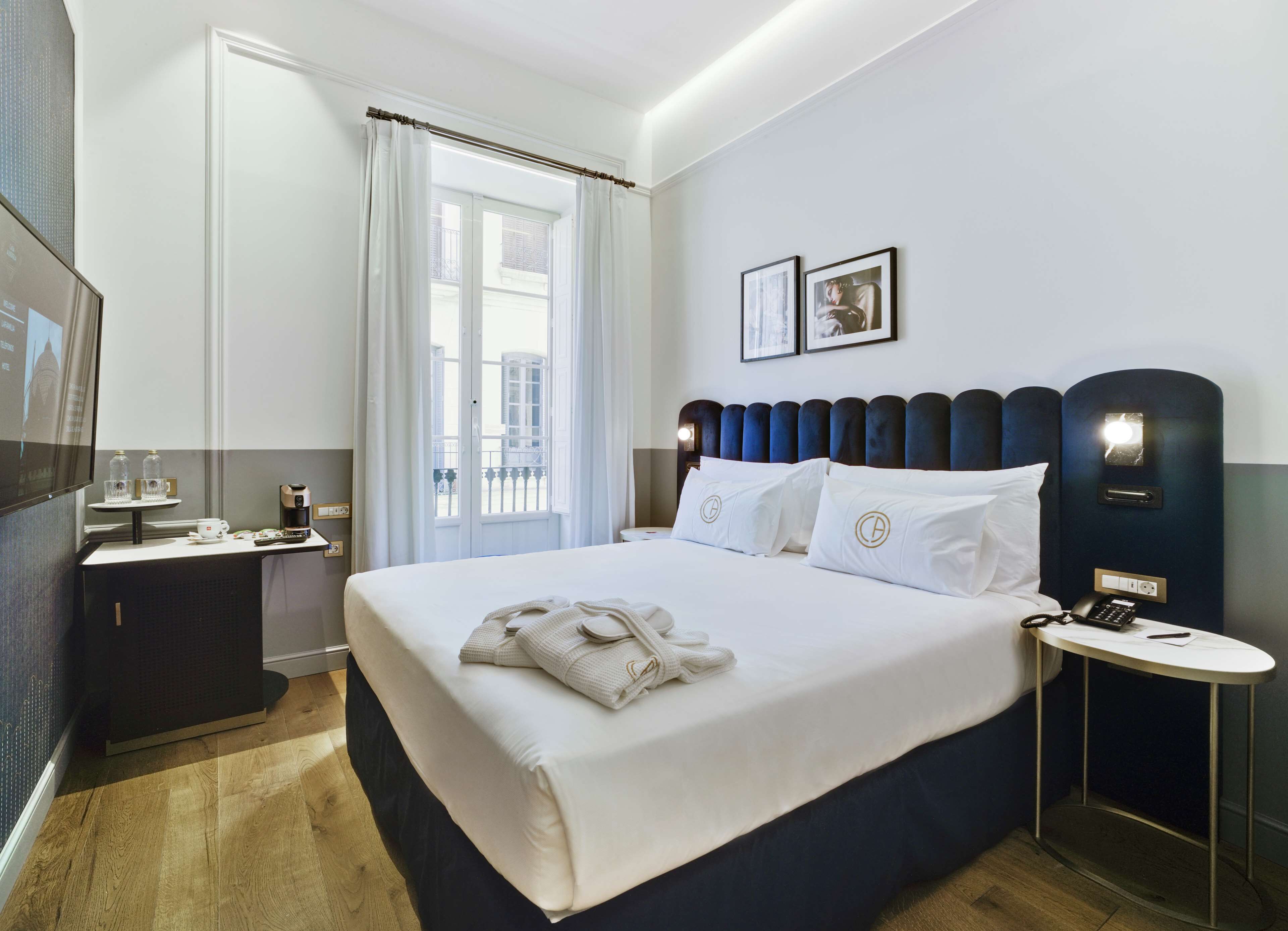 A hotel room with a large bed, a bedside table with a phone, a small table with a coffee maker, and a window with curtains.