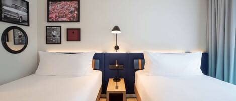 Habitación de hotel con dos camas, una mesita de noche con lámpara y cuadros en la pared.