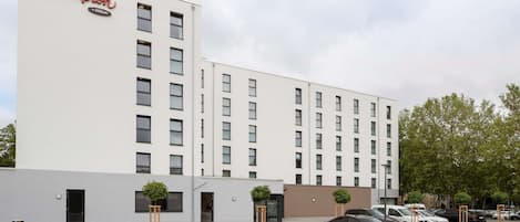 A modern hotel building with a prominent sign, a parking lot, and several cars parked.