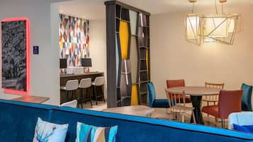 Una hall d'hotel moderna con un'area salotto blu, un tavolo da pranzo centrale con sedie, un'opera d'arte geometrica a parete e un lampadario particolare.