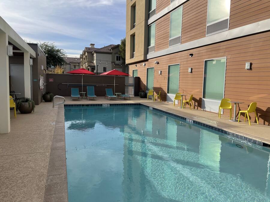 Una piscina con sillas de descanso y sombrillas, rodeada de edificios con amplias ventanas y una terraza con baldosas.