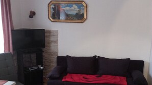 Una sala de estar con un sofá negro, una manta roja, un televisor sobre un mueble bajo y una chimenea.