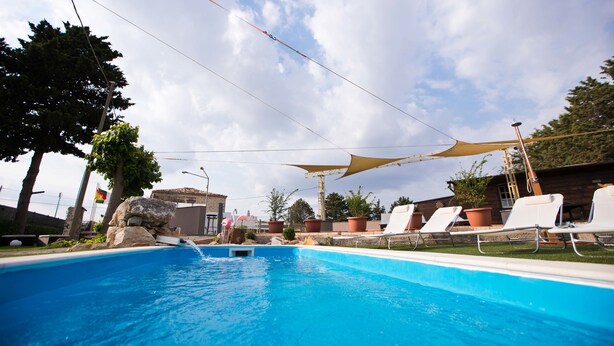 Una piscina con sillas de descanso y una zona sombreada, rodeada de árboles y plantas en macetas.