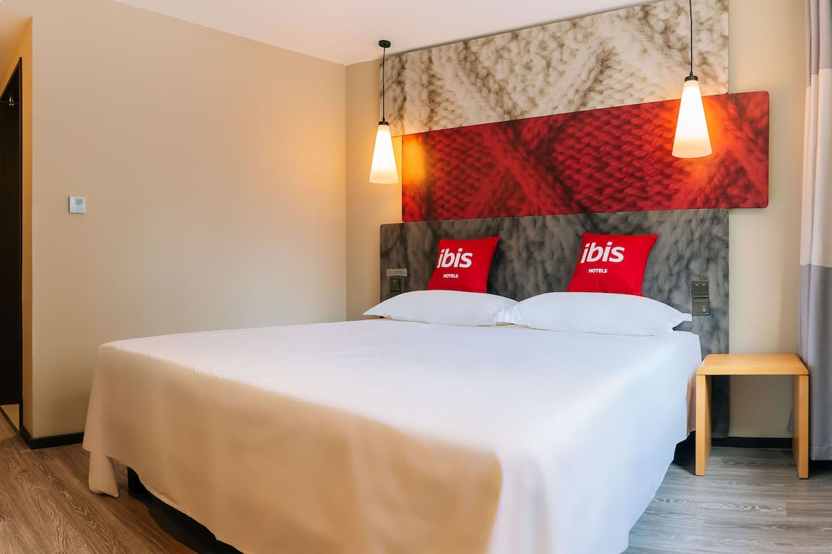 Habitación de hotel con una cama grande, dos almohadas rojas, un taburete de madera y una lámpara fijada a la pared.