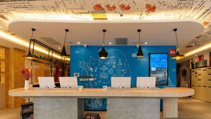 Ein Empfangsschalter mit zwei Laptops, eine blaue Wand mit weißen Kritzeleien und ein Schild mit der Aufschrift 'ibis'.