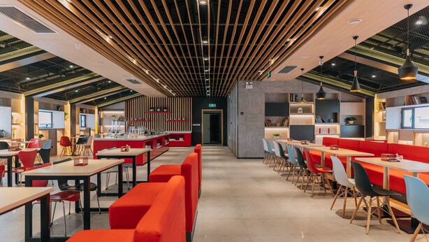 Ein modernes Restaurant mit hohen Decken, freiliegenden Balken und einer Mischung aus roten und blauen Sitzgelegenheiten.