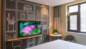 Ein modernes Hotelzimmer mit einem großen Flachbildfernseher an der Wand, einem kleinen runden Tisch mit einer Pflanze, einem bequemen Sessel und einem Fenster mit Blick auf Gebäude.