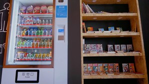 Una máquina expendedora llena de bebidas y aperitivos, junto a una estantería de madera con libros y adornos.