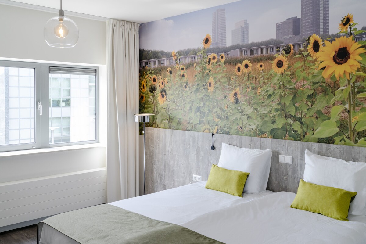 A hotel room with a large bed, two pillows, a cityscape mural on the wall, and a window with curtains.