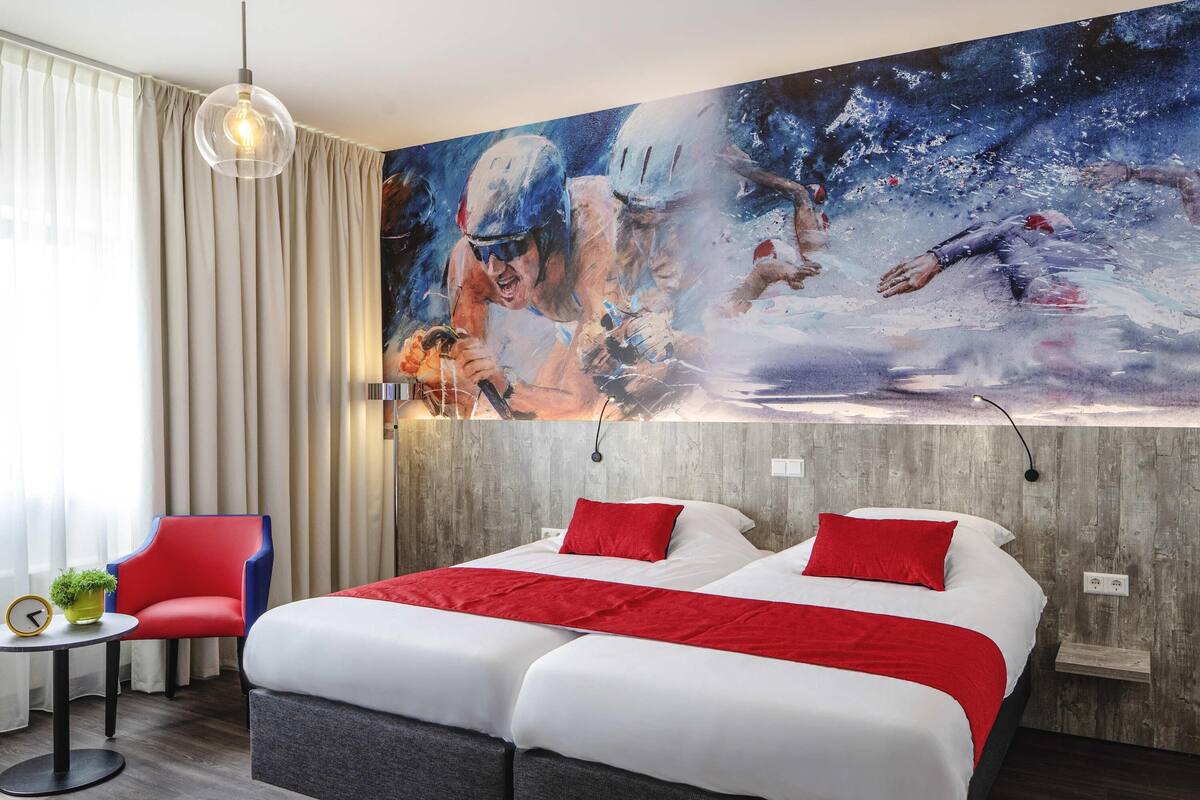 Una habitación de hotel con una cama grande, ropa de cama roja y blanca, una silla roja, una mesita con una planta y un cuadro de nadadores en la pared.