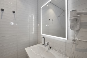 A modern bathroom with white tiled walls, a large mirror, a sink, and a hairdryer mounted on the wall. - ibis Styles Almere (Almere)