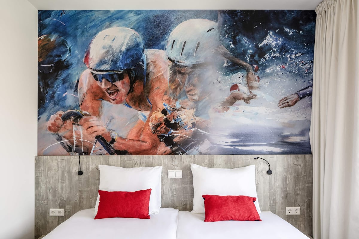 Habitación de hotel con un cuadro abstracto grande en la pared, un cabecero de madera y dos camas con almohadas rojas y blancas.