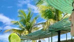 Eine Person hält ein Getränk mit einer Limettenscheibe, neben einem Pool mit Palmen und einem Holzzaun im Hintergrund.