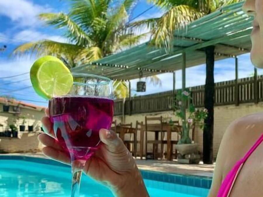 A person holding a drink with a lime slice, by a pool with palm trees and a wooden fence in the background.
