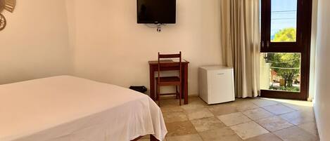 Une chambre d’hôtel comprenant un lit, un bureau avec une chaise, une télévision, un réfrigérateur et une porte donnant sur un balcon.