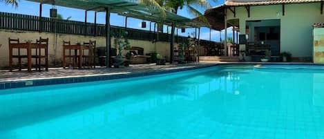 Una piscina cristalina con bordes de azulejos, rodeada por un área de estar techada con sillas y mesas de madera, palmeras y un edificio con techo de tejas.