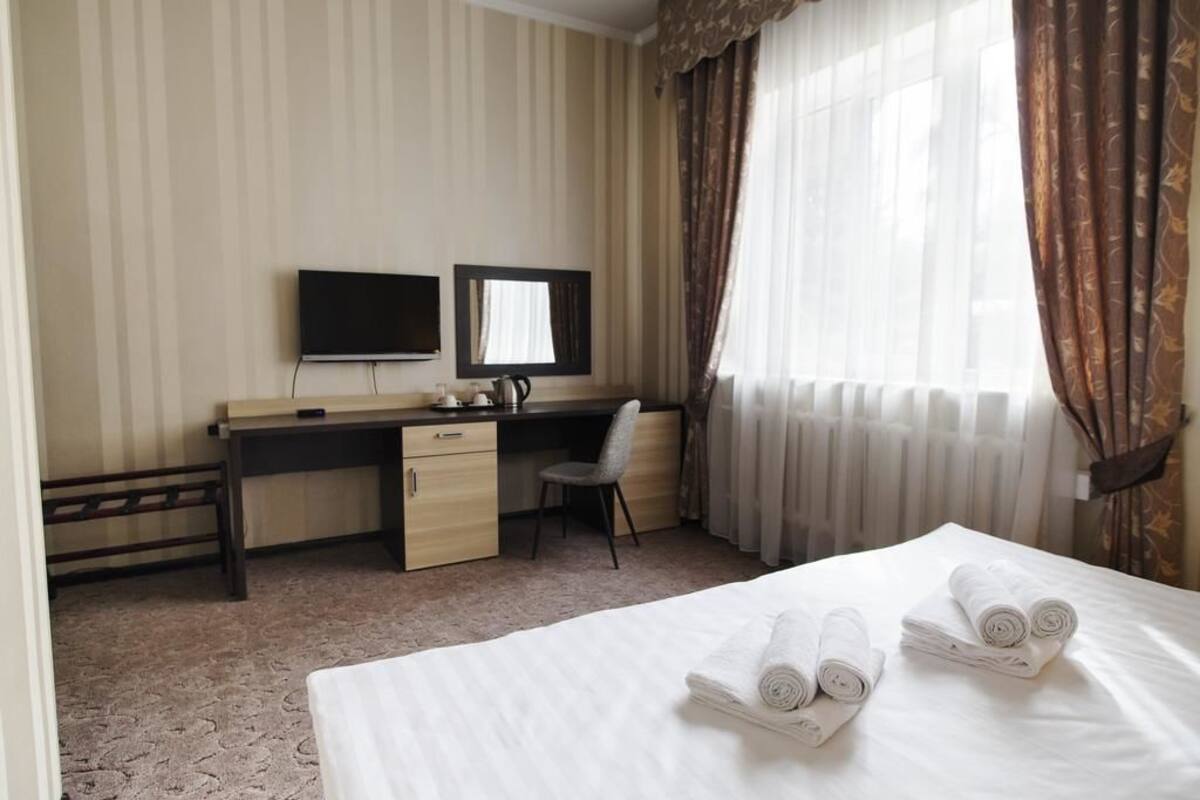 A hotel room with a bed, a desk with a chair, a television, and a window with curtains.