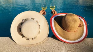 Deux personnes, chacune tenant un verre de vin, sont assises au bord d’une piscine. Un grand chapeau et un plus petit chapeau sont posés à côté d’elles.