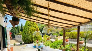 Un espace de restauration en plein air avec une table en bois prévue pour quatre personnes, entourée de plantes en pot et surmontée d’une pergola.