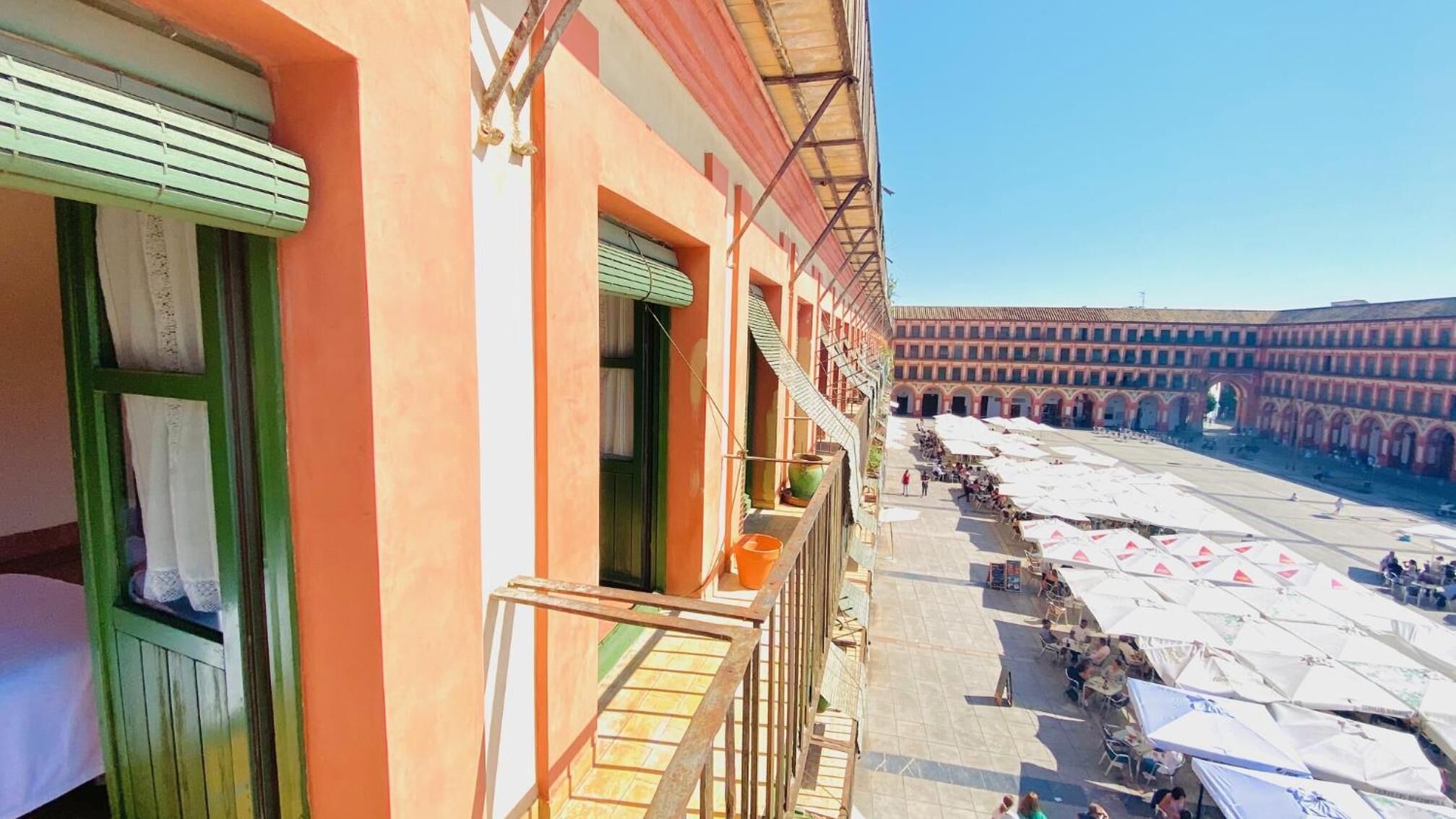 A building with a balcony overlooking a courtyard with outdoor seating and a large arched structure in the background.