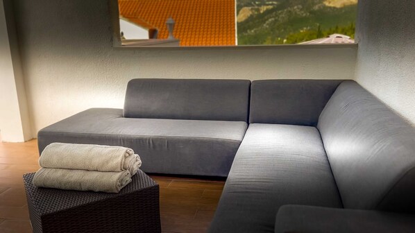 Un sofá gris con una manta doblada sobre un reposapiés de mimbre en una habitación con un ventanal grande.